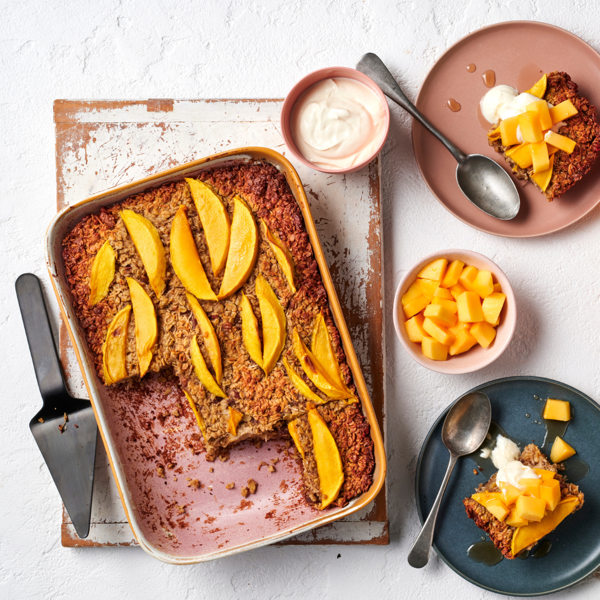 A birdseye view of a baking dish with mango baked oatmeal, with several slices cut out and shown on two plates nearby