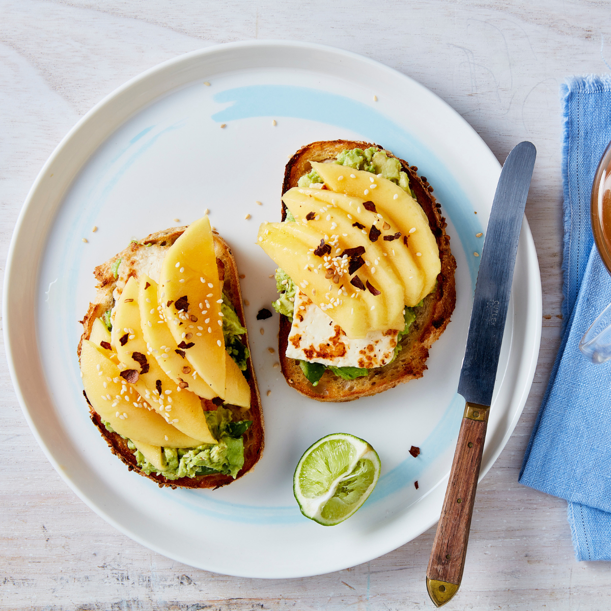 two pieces of sourdough toast on a white plate topped with avaocado, haloumi, mango, chilli flakes and sesame flakes. The background is white rustic wood