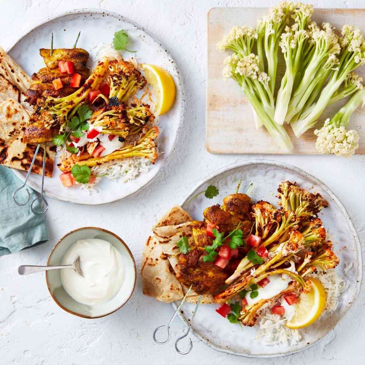 Two plates with tandoori roasted cauliblossom and BBQ chicken skewers topped with yoghurts, tomatoes and lemon. 